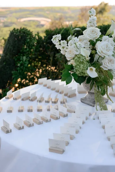 Escort card display with wood place card holders and white roses, arranged outdoors amid garden greenery, trimmed hedges, and distant hills.