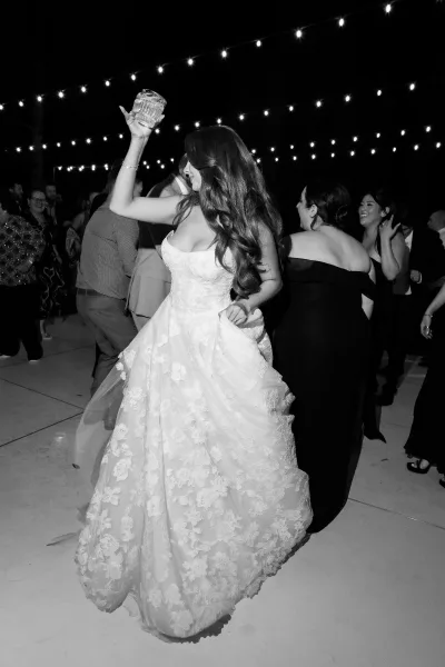 Bride dancing on the reception dance floor, raising a cocktail glass with a joyful grin beneath the night sky, motion captured mid-twirl.