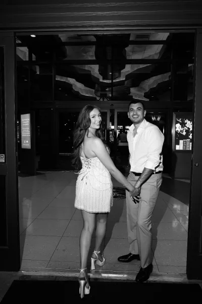 Couple portrait of a bride and groom holding hands, she in a short bridal dress, posed by glass doors on a tiled lobby floor as they look back.