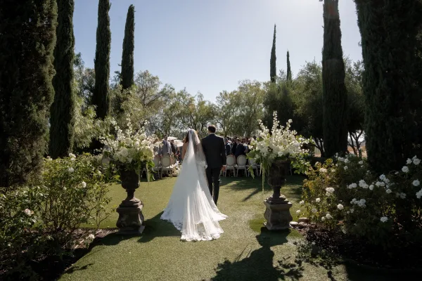 Ceremony processional with the bride walking down the aisle, long veil trailing, framed by cypress trees under a clear blue sky.