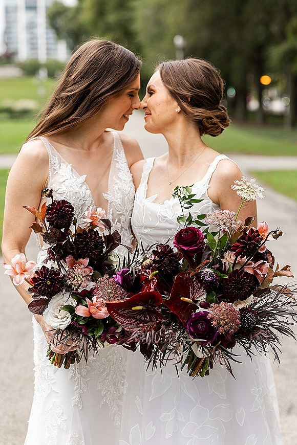 Bride couple portrait of two brides nose to nose, holding dahlia and calla lily bouquets on a park path with trees and streetlights behind