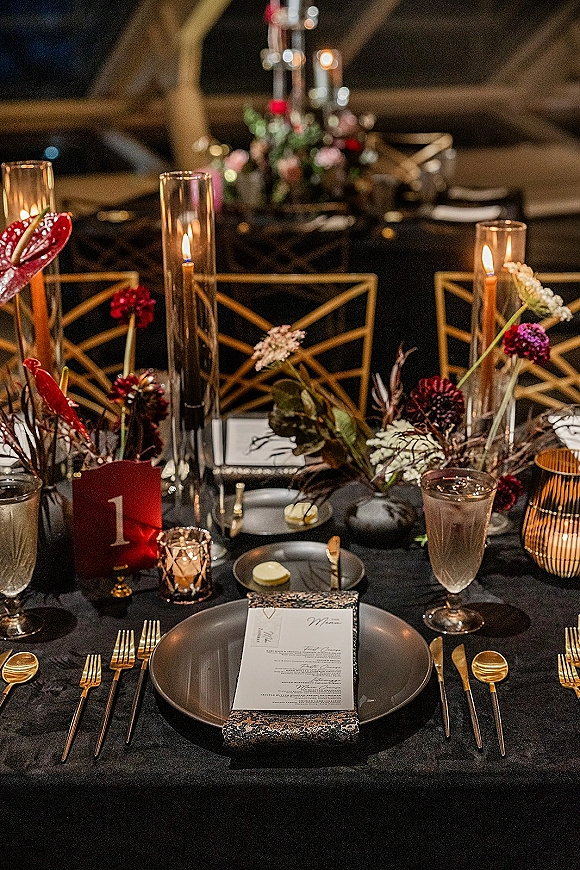 Reception tablescape with black wedding tablescape details, gold flatware, black plates, glass goblets, taper candles, and anthurium florals under dim lighting