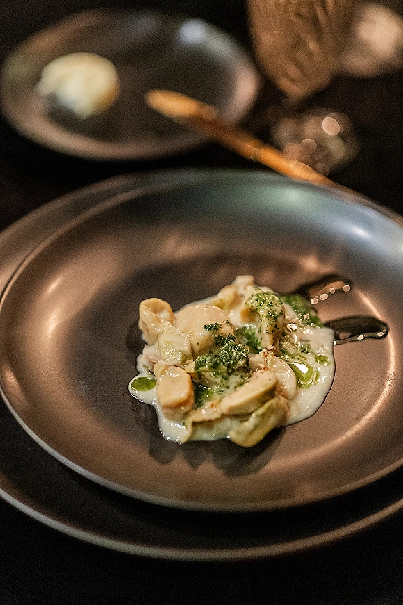 Wedding reception meal on ceramic plates with pesto sauce, spoon, bread roll, and wine glass arranged on a dark tabletop place setting
