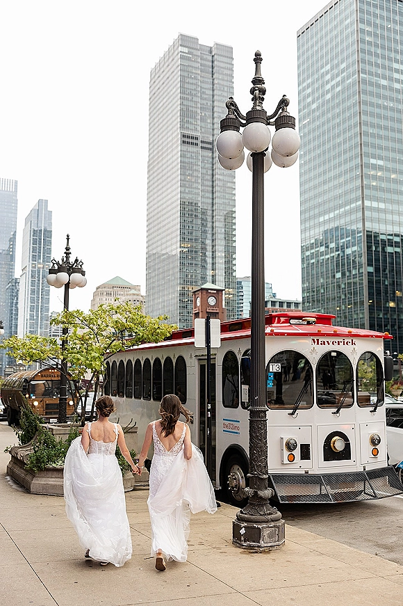 Bridal party walking hand in hand, bridesmaids walking away in flowing tulle skirts along a city street beside a trolley bus