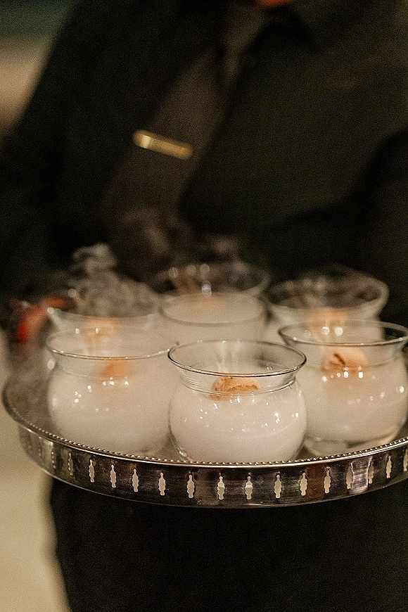 Wedding signature drink smoke bubble cocktail served in clear glasses on a metal tray, held by a server in a dark suit under warm lighting