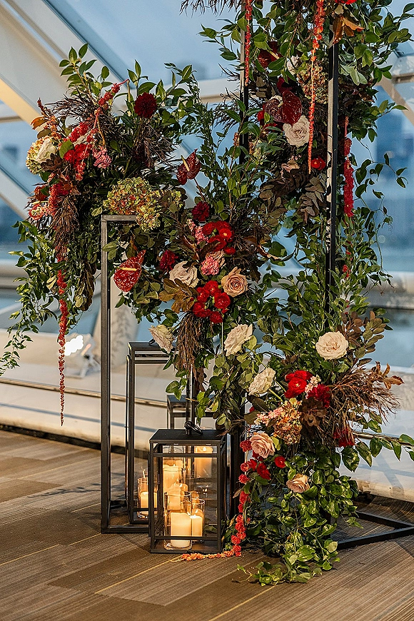 Wedding altar decor with an asymmetrical wedding arch on a black metal frame, blush and red blooms, greenery garland, and lanterns in an indoor venue