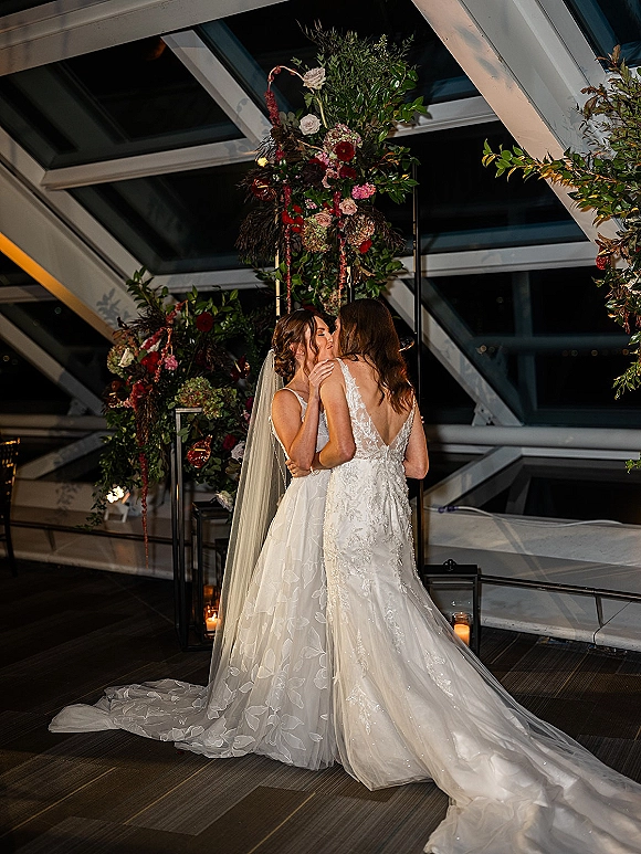 Wedding kiss between two brides beneath a floral arch with greenery and lanterns, cathedral veil flowing in a windowed industrial venue at night