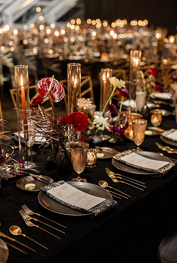 Reception tablescape with a black wedding tablescape, taper candles and glass hurricanes glowing over gold flatware and anthurium florals under string lights