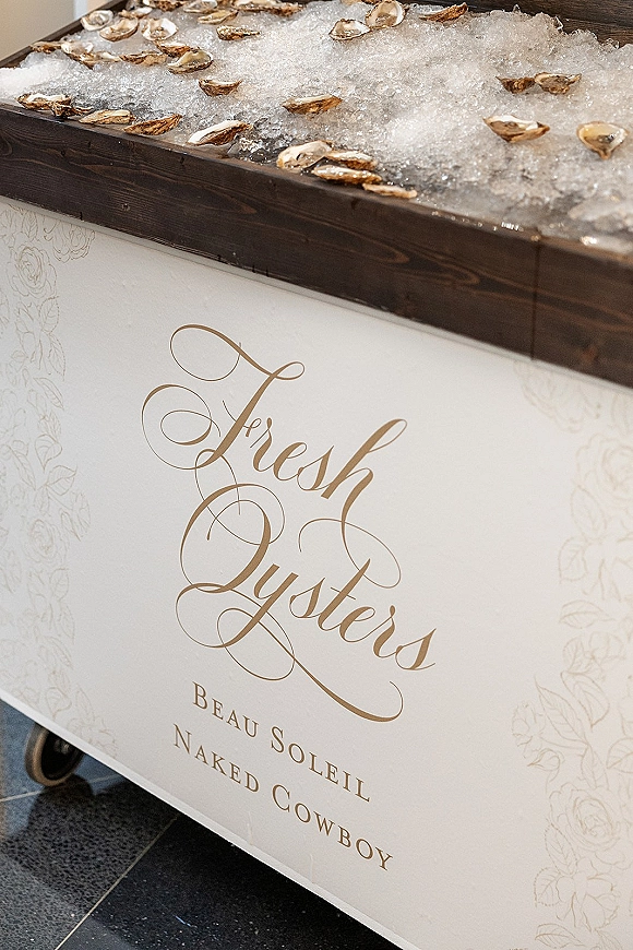 Oyster bar with wedding oyster bar sign, oysters on the half shell on crushed ice atop a wooden tray on a white cart with floral panel