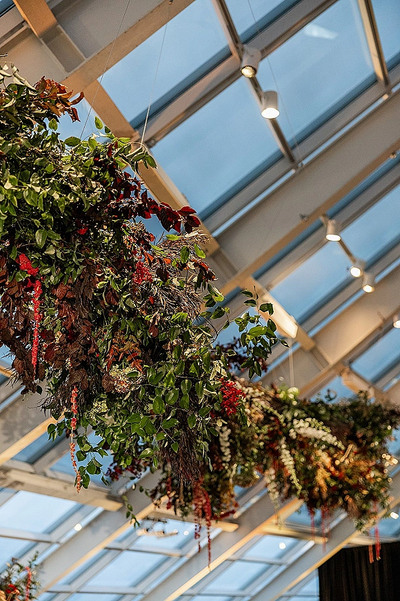 Hanging floral installation with ceiling greenery and red flowers cascading beneath skylight panels, beams, and pendant lights overhead