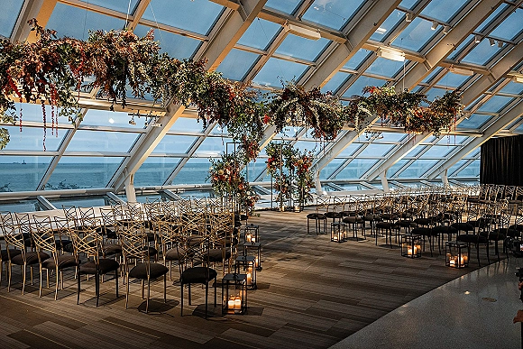 Ceremony setup for an indoor wedding ceremony with gold chairs and candlelit aisle, beneath hanging florals by ocean-view windows under glass ceiling