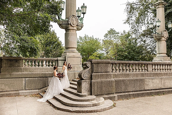 Bride portrait on stone staircase holding a burgundy bouquet, her wedding dress train draped by a classic balustrade and trees beyond