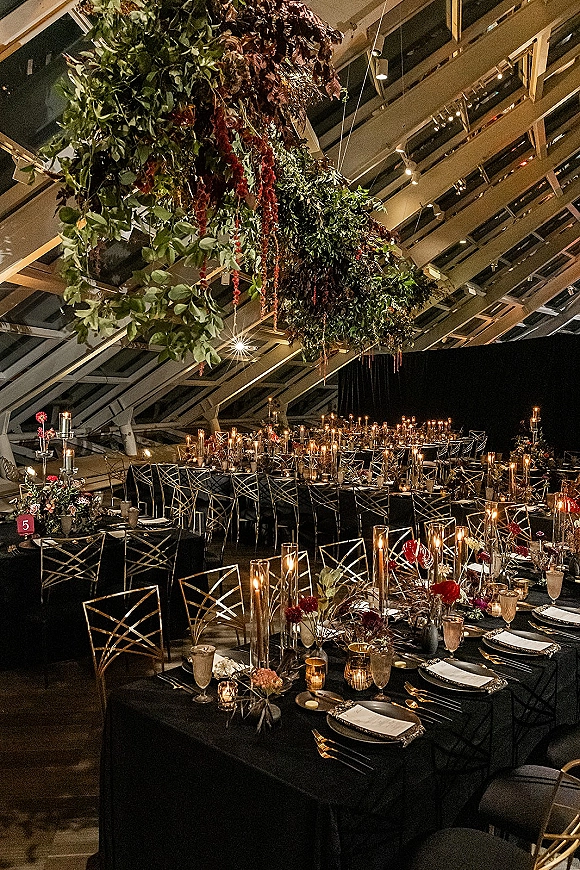 Reception tablescape with black wedding tablescape details, gold chairs, candlelit florals, and hanging blooms under string lights in an industrial space