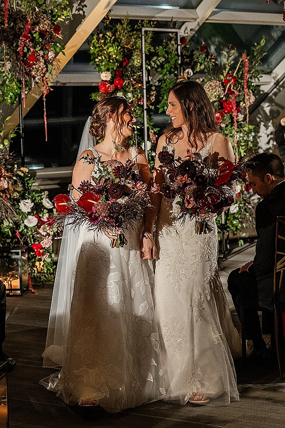 Ceremony moment at a same sex wedding ceremony as two brides hold hands in wedding dresses beneath a greenery arch, lanterns glowing indoors