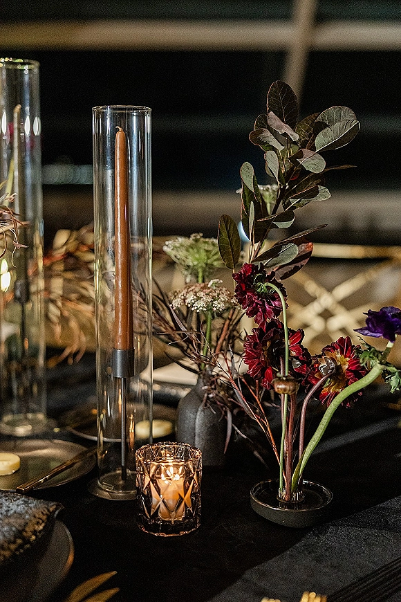 Reception tablescape with a moody wedding tablescape of taper candles, glass hurricanes, and floral greenery on black linens in a dark interior