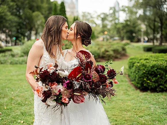 Wedding kiss portrait of two brides kissing in lace dresses, holding a burgundy anthurium bouquet on a park lawn with trees and city bokeh