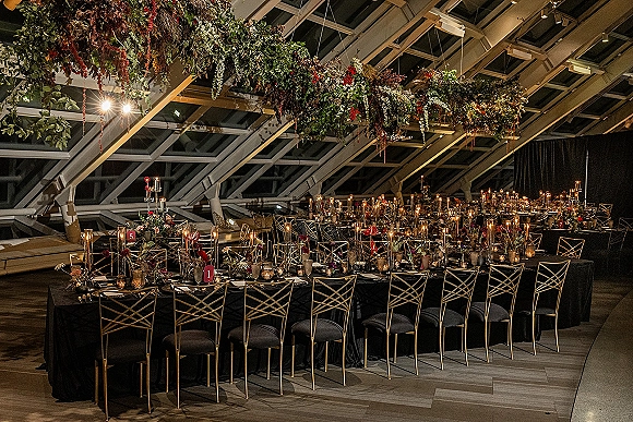 Reception tablescape with long banquet table setup in black linens, gold chairs, tall taper candles and florals beneath hanging greenery and glass ceiling