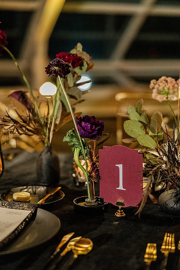 Reception tablescape with a wedding table number card in a gold holder, bud vase florals, taper candles, and black-and-gold place settings in a dim room