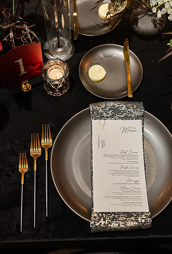 Reception tablescape with black tablecloth tablescape styling, gold flatware, gray plates, patterned napkin, menu card, votive and florals on dark table