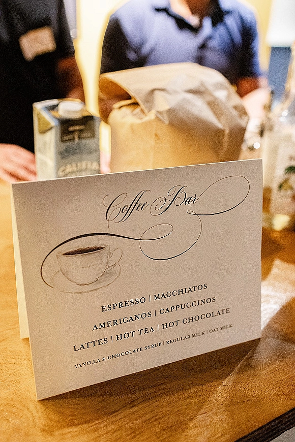 Coffee bar sign with calligraphy menu and coffee cup illustration on a wood table beside cartons and a glass dispenser at reception