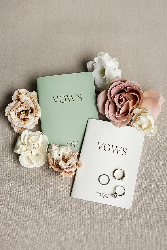 Vow books with gold foil lettering beside wedding rings and flowers, styled in a minimalist flat lay on a linen fabric backdrop
