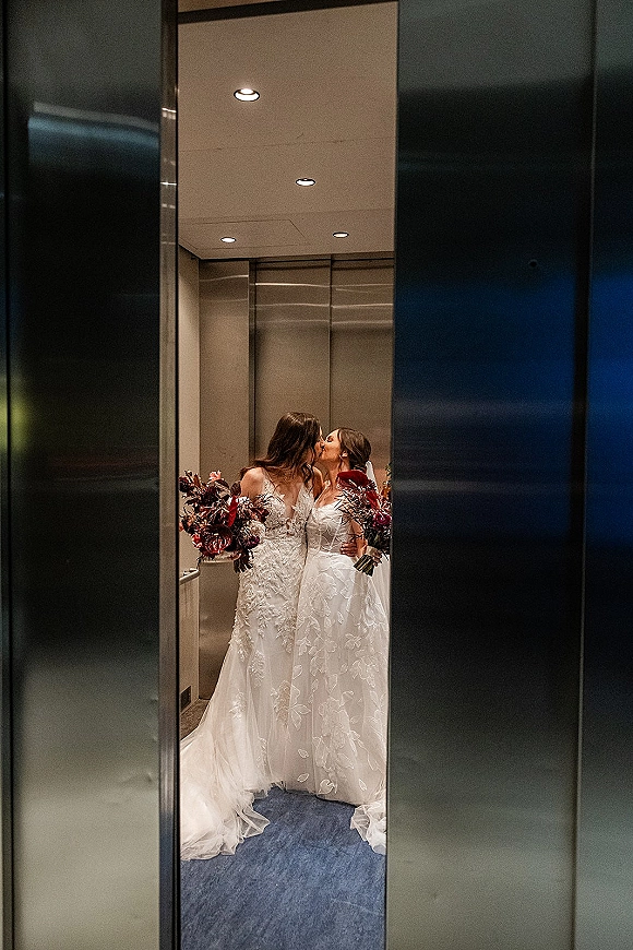 Wedding kiss portrait of two brides kissing in a metal-walled elevator, holding burgundy bouquets in lace gowns with a veil and long train