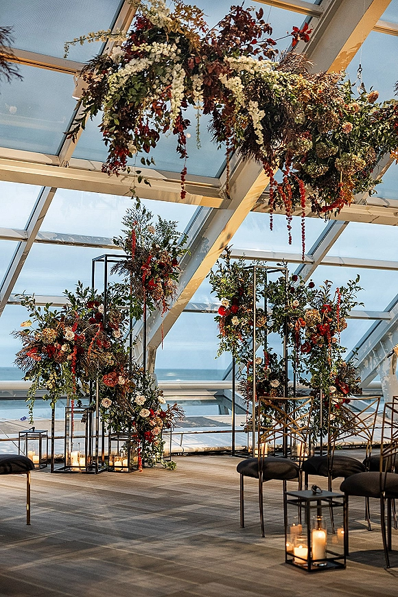 Wedding ceremony decor with hanging floral installation and greenery on black metal frames, accented by pillar candles under a glass ceiling with ocean view