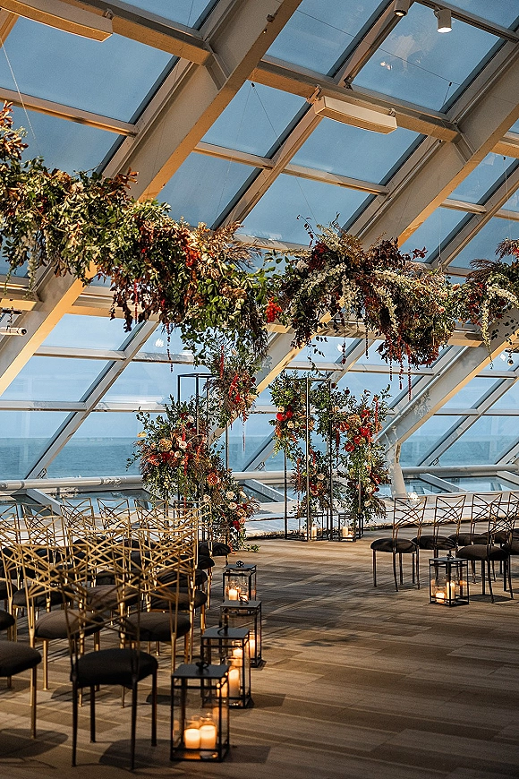 Ceremony setup with hanging greenery installation above a candle-lined aisle, framed arches and mixed chairs in a glass venue with ocean view