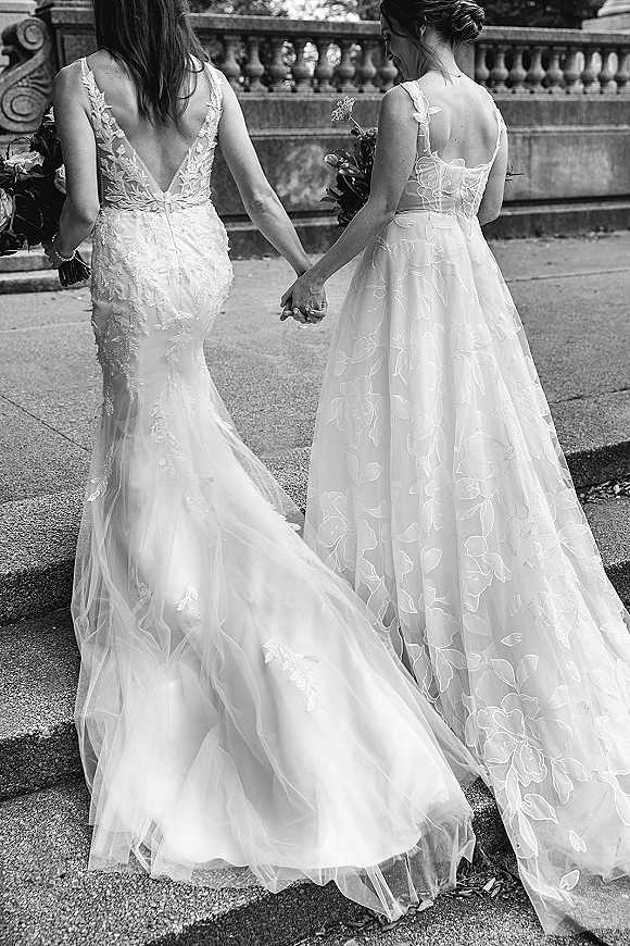 Brides holding hands, same sex wedding brides walking away in lace and tulle gowns with bouquets along a stone balustrade walkway