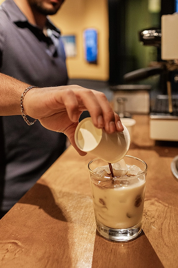 Iced coffee pour into a glass tumbler iced latte with ice cubes as cream swirls in, on a wood coffee bar counter with wall art