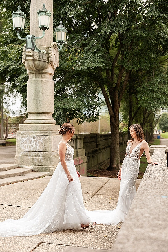Bride portrait in a lace wedding dress, looking down while leaning on a stone bridge railing with vintage street lamps and trees behind