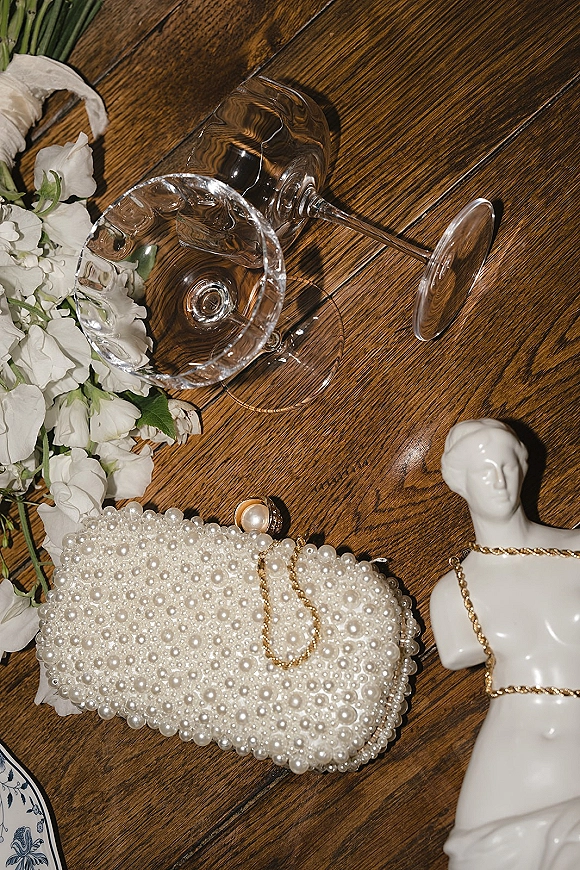 Wedding accessories flatlay featuring a pearl clutch bag with gold chain necklace, pearl earrings, champagne coupe glasses, and white flowers on a wood table