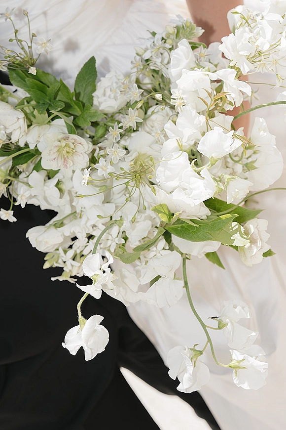 Bridal bouquet of white wedding bouquet blooms with lush greenery, trailing stems and ribbon, held beside a black suit jacket and bridal gown