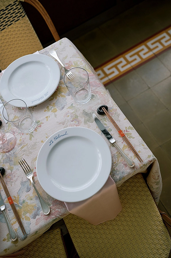 Reception tablescape with a floral tablecloth, white plates, blush napkins, flatware, wine and water glasses, and chopsticks on woven chairs by banquette seating