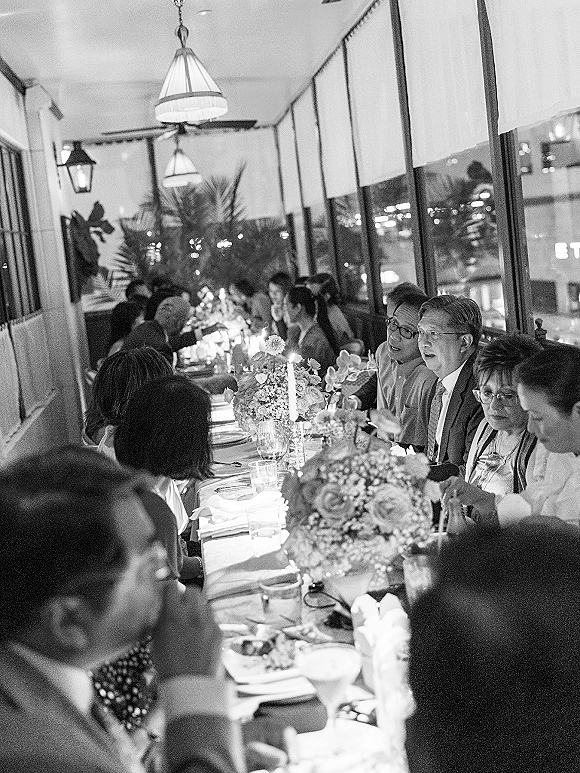 Wedding reception dinner at a long banquet table with floral centerpieces and taper candles in a restaurant with city lights outside windows
