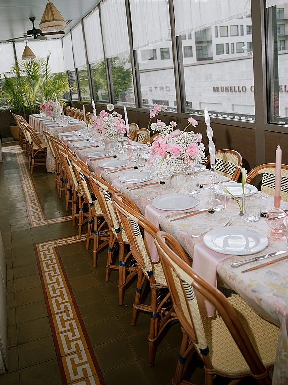Reception tablescape with a long banquet table wedding setup, pink runner, taper candles, floral centerpieces, and rattan chairs by window wall