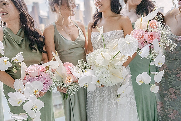 Bridesmaid group portrait with bride and bridesmaids in sage green dresses holding blush and white bouquets on a terrace with city skyline backdrop