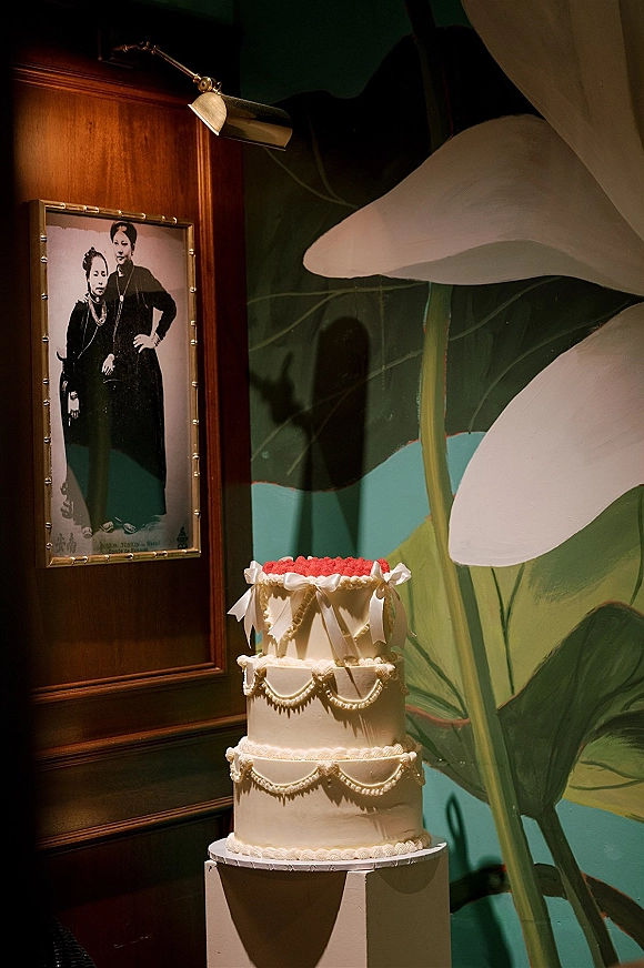 Wedding cake with piped frosting on a pedestal plinth, finished with white ribbon bows and a red rosette topper before a floral mural wall