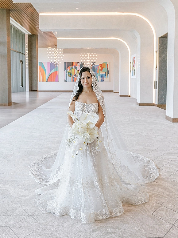 Bridal portrait of a bride holding bouquet in a strapless lace wedding dress and cathedral veil, standing in a modern hotel lobby under chandeliers