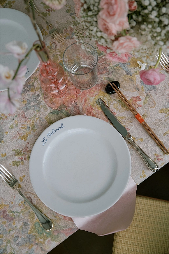 Reception place setting with white plate, pink napkin, silver cutlery, pink goblet, chopsticks, and calligraphy place card on floral tablecloth