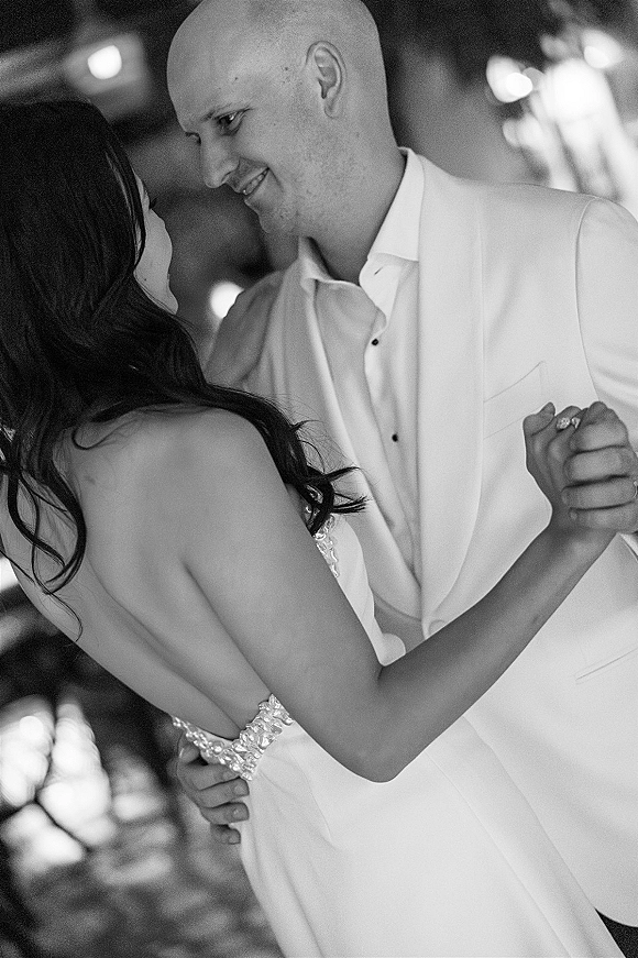 First dance as bride in a beaded strapless wedding dress and groom in a white suit hold close in an indoor reception with blurred lights