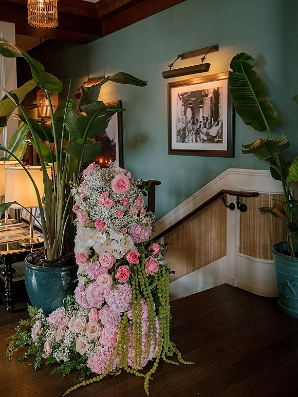 Wedding floral arrangement of pink roses, hydrangea and white orchids with cascading amaranthus beside a teal-walled staircase indoors