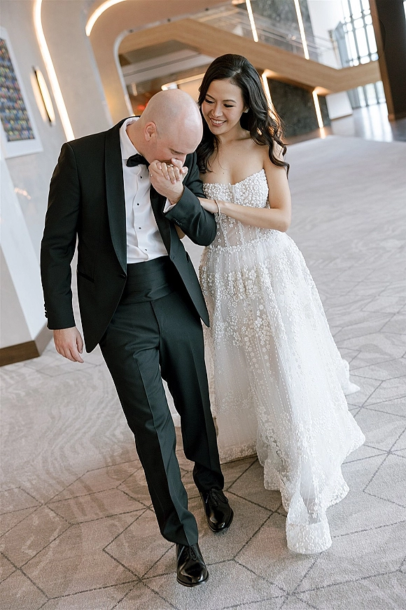 Couple portrait of groom kissing bride’s hand as they walk arm-in-arm, her strapless lace gown and his bow tie in a modern arched venue