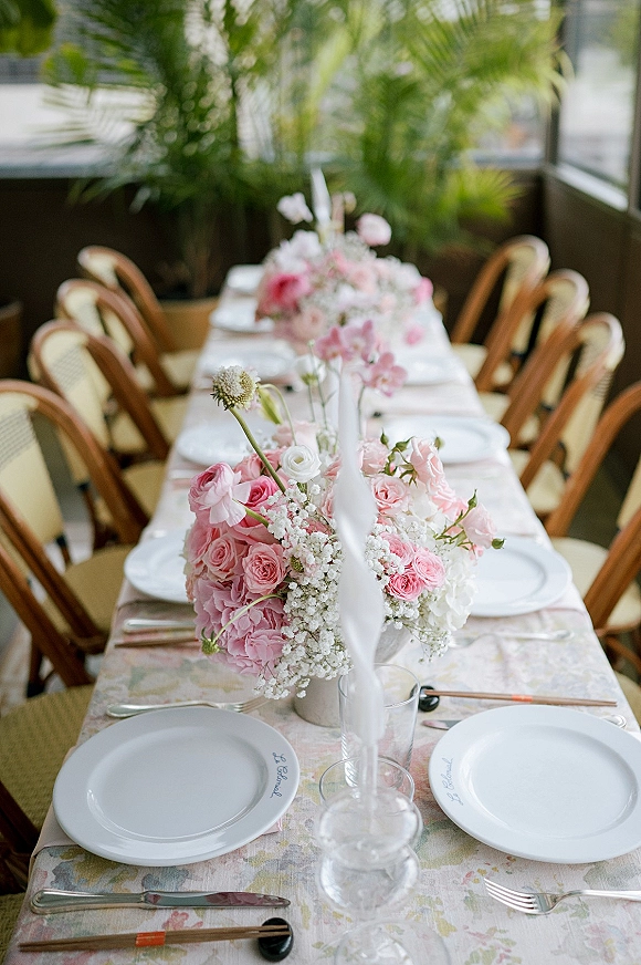 Reception tablescape with pink wedding centerpieces, white taper candles, floral tablecloth, and rattan bistro chairs in a bright indoor dining area