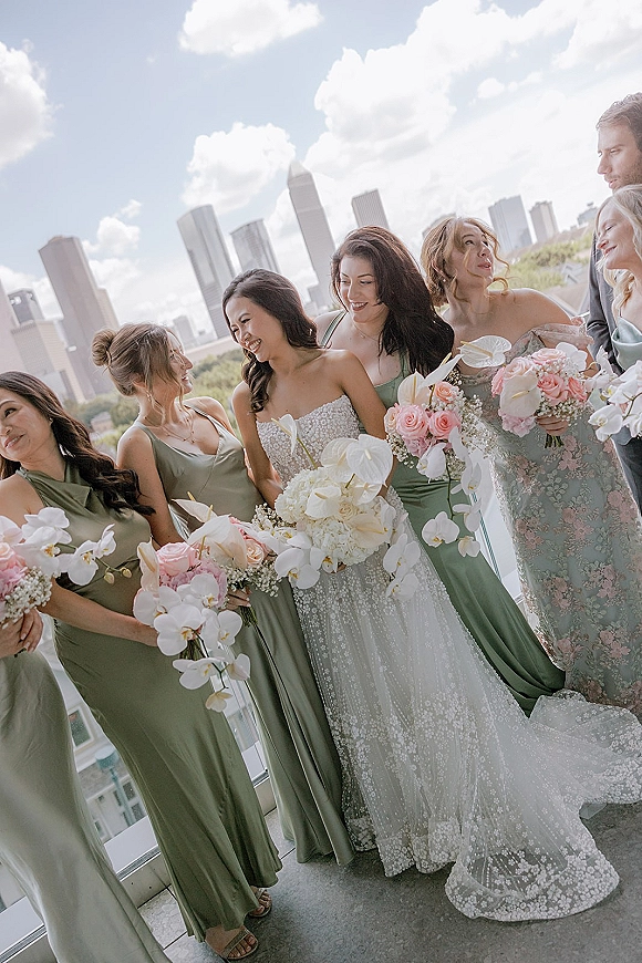 Bridesmaid group photo with bride and bridesmaids holding pink and white bouquets on a balcony, city skyline behind them