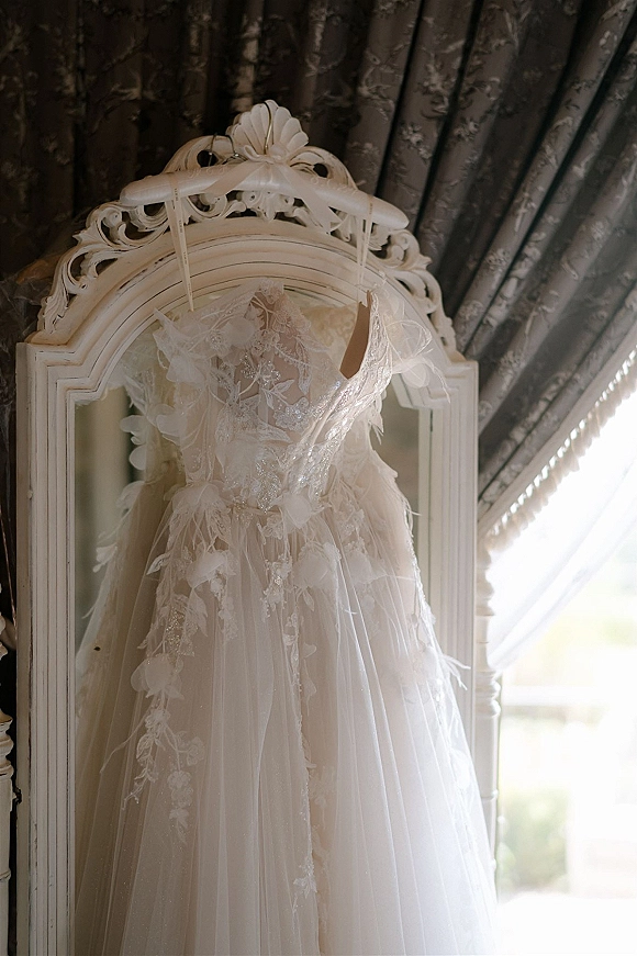 Wedding dress with lace wedding dress appliqué and beaded bodice on a satin ribbon hanger beside an ornate mirror in window light