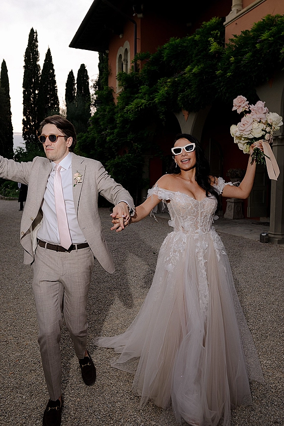 Newlywed exit as bride and groom walk hand in hand, bride lifting bouquet, wearing sunglasses in an ivy-covered villa courtyard