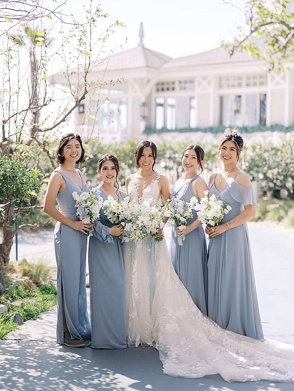 Bridesmaids portrait with bride in a lace gown, women in dusty blue dresses holding white and green bouquets on a garden walkway near a gazebo