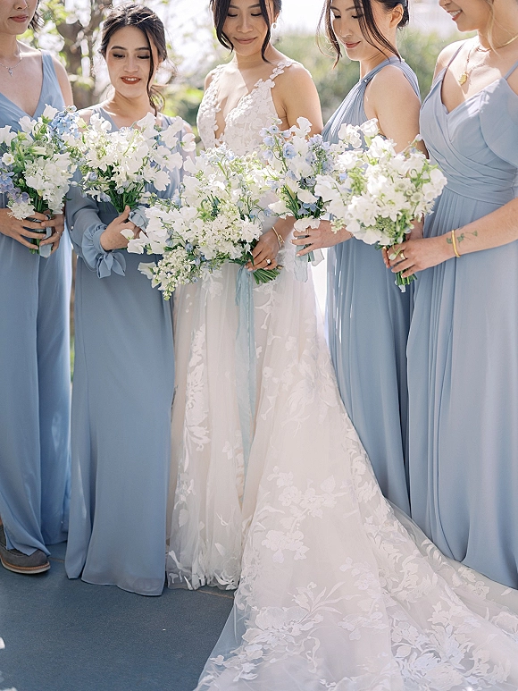 Bride with bridesmaids in blue bridesmaid dresses holding white and blue bouquets outdoors amid lush greenery in soft daylight