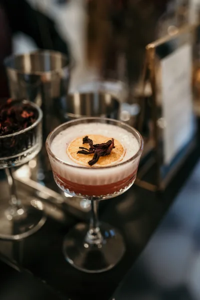 Wedding signature cocktail in a coupe glass with signature drink garnish of citrus and dried flower on a bar counter in a blurred reception setting